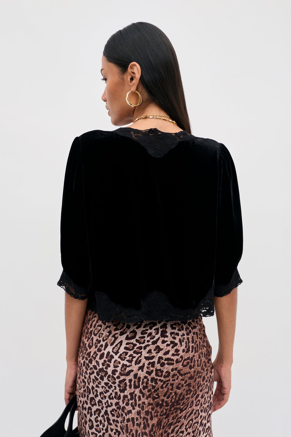 A woman with long dark hair stands facing away, wearing the Ursula - Black, a vintage party top in black silk velvet with delicate lace trim, paired with a leopard print skirt. She accessorizes with large hoop earrings and holds a small black bag against a plain, light-colored background.