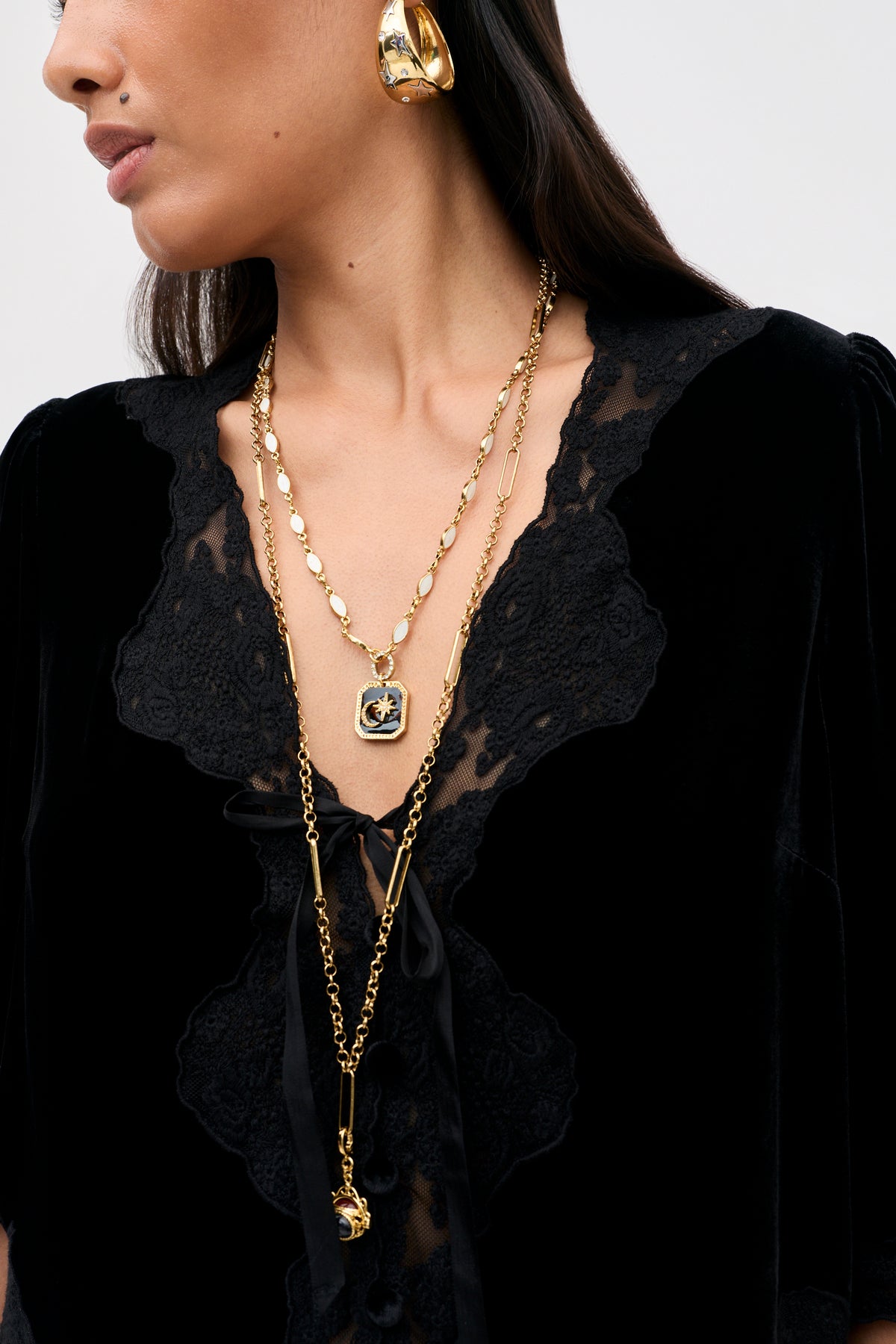A woman wears the Ursula - Black vintage party top with detailed lace trim, paired with large gold hoop earrings and two layered gold necklaces featuring decorative pendants. The focus is on her jewelry and the intricate texture of the Ursula - Black top.