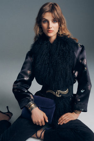 A person wearing a Kove - Black embroidered jacket with a fur collar, a gold snake-shaped belt, and large gold earrings, holding a dark purple clutch.