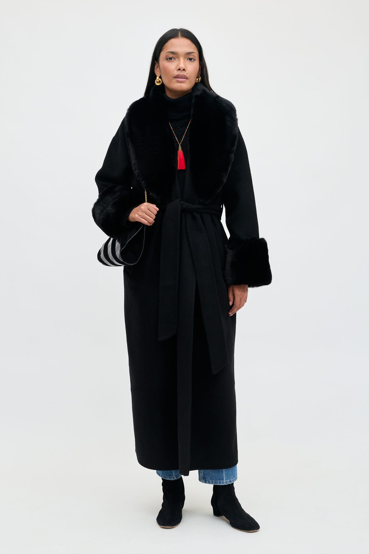 A woman wears a Winslow - Black long wrap coat with faux fur collar and cuffs, a red tassel necklace, blue jeans, and black ankle boots while carrying a striped shoulder bag.