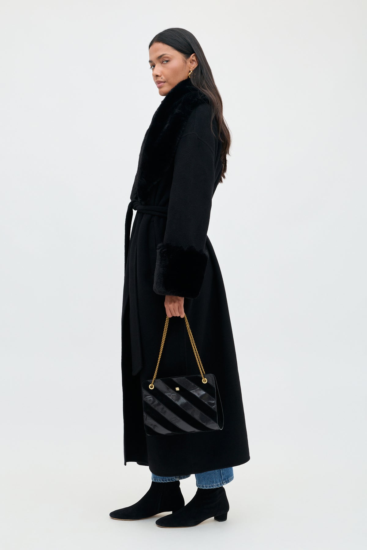 A person wearing the Winslow - Black, a long coat with faux fur trim on the collar and cuffs, holding a black quilted handbag with a gold chain strap.