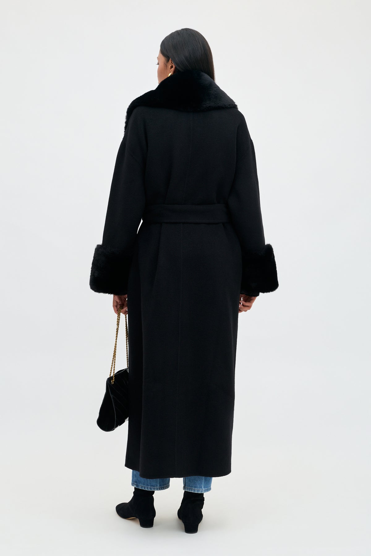 A person seen from behind wears the Winslow - Black coat with a faux fur collar and cuffs, jeans, and ankle boots, while holding a small black shoulder bag.