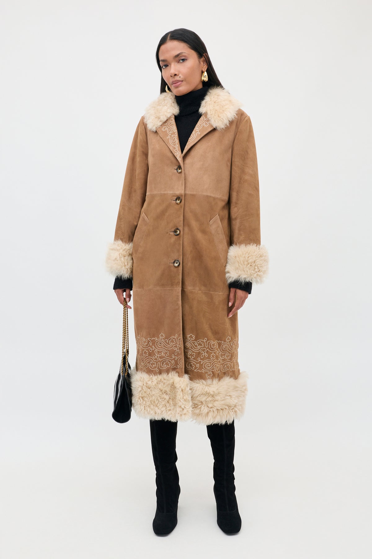 A woman wearing the Cienna - Camel shearling coat with fur trim, paired with a black turtleneck, black pants, and boots. She holds a black handbag and stands against a plain white background, highlighting the chic appeal of this classic Cienna - Camel coat.