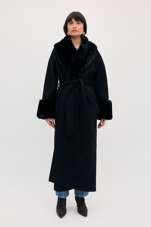 A person wearing a Winslow - Black belted coat with faux fur trim on the collar and cuffs over jeans and heeled boots.