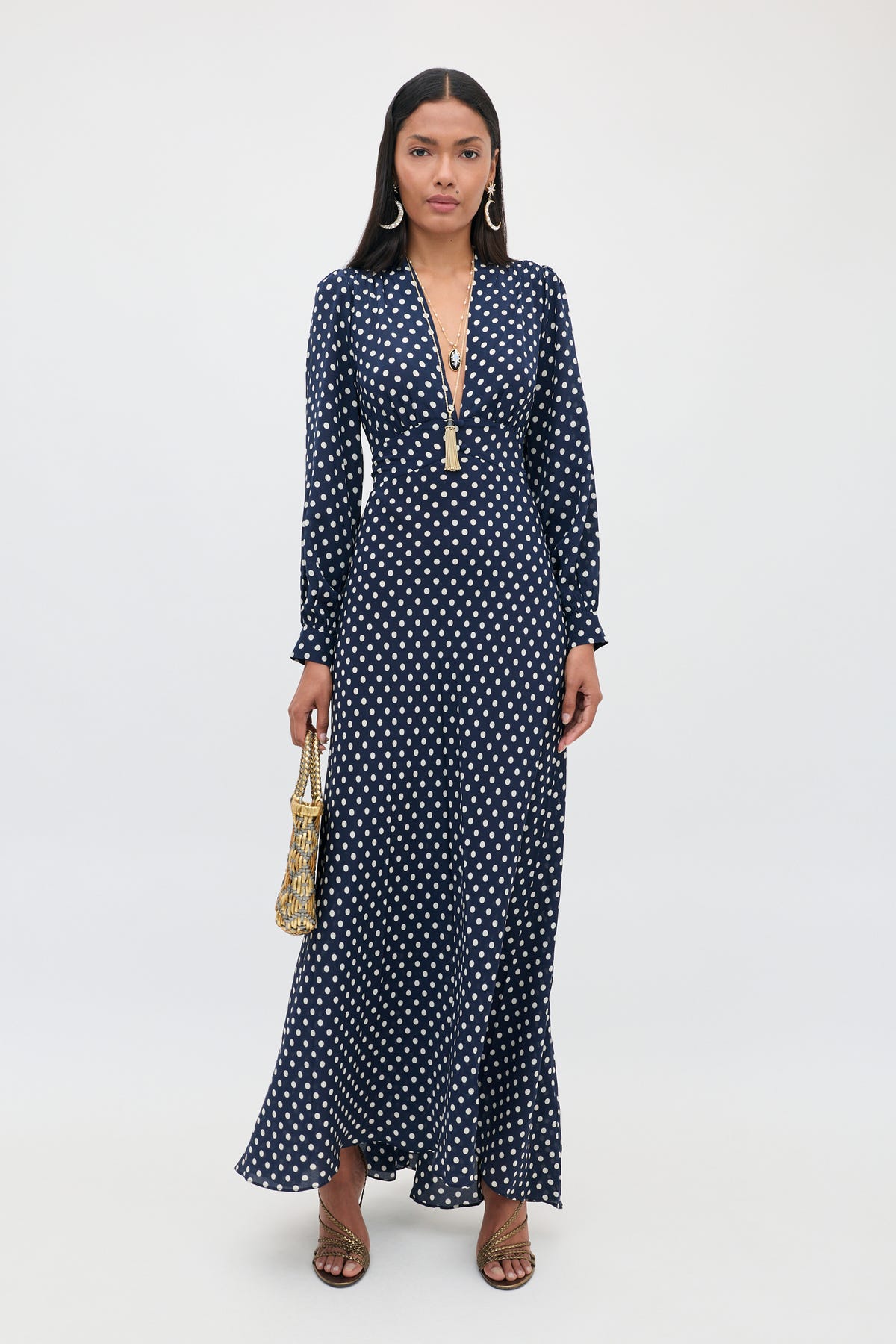 A woman stands against a plain white background wearing the Emory - Runway Spot Navy dress, which features long sleeves, a deep V-neck, and navy blue polka dots; she accessorizes with a gold handbag, long earrings, and sandals.