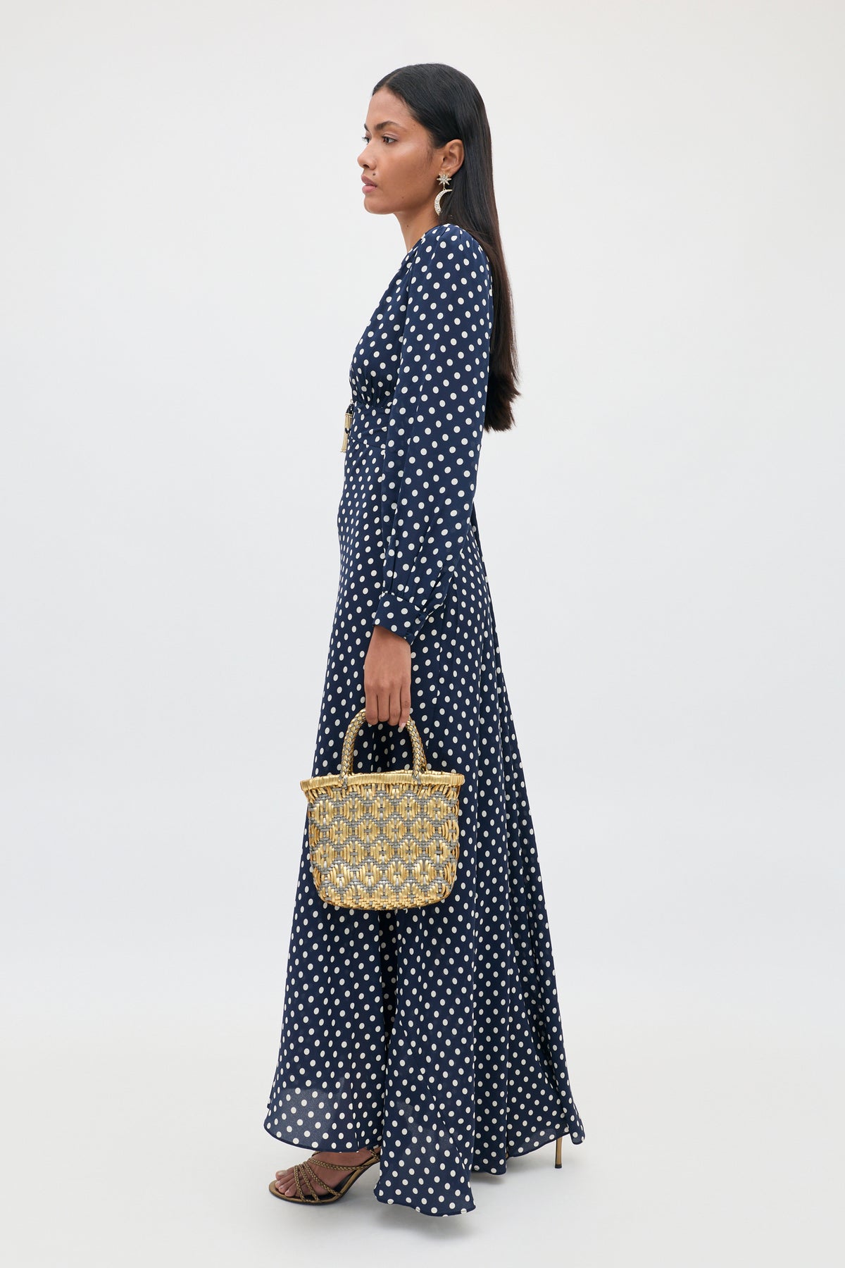 Woman in a navy blue, long-sleeved Emory - Runway Spot Navy dress adorned with chic polka dots, holding a woven handbag, wearing heeled sandals and statement earrings, standing against a plain light background.