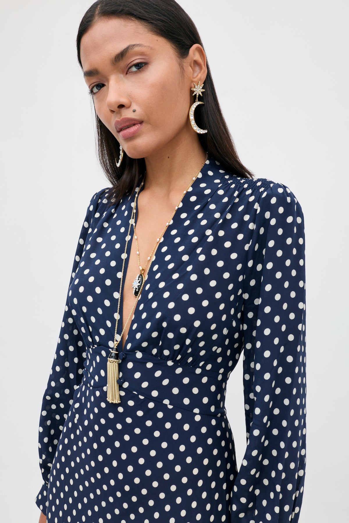 A woman wears the Emory - Runway Spot Navy dress, featuring navy polka dots, long sleeves, and a deep V-neck. She accessorizes with statement jewelry—a pair of dangling earrings and a necklace—and has long dark hair and a neutral expression.