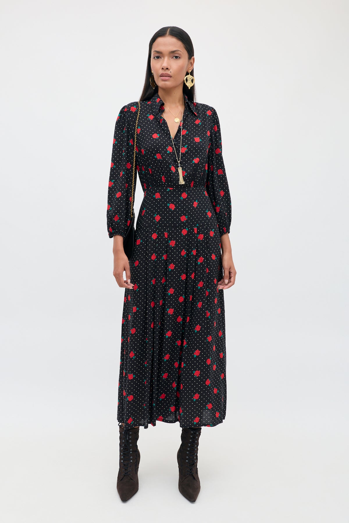 A woman wearing the “Izzy - Rose Spot Black” dress, featuring a long black silhouette with red polka dots, long sleeves, and a V-neckline. She has paired the dress with tall brown lace-up boots and has long dark hair, standing against a plain white background.