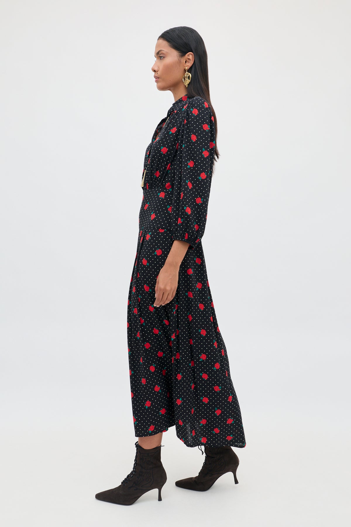A woman stands in profile wearing the Izzy - Rose Spot Black, a long-sleeve black dress adorned with red polka dots. She pairs the dress with black lace-up ankle boots and large earrings, set against a simple, light-colored background.