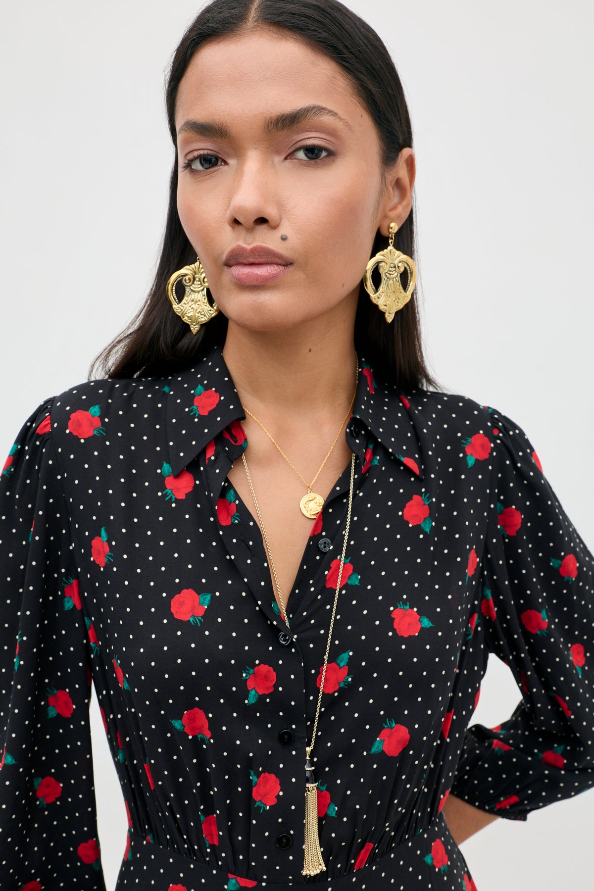 A woman wears the Izzy - Rose Spot Black dress, featuring red roses and white polka dots, paired with gold earrings and layered necklaces, standing against a plain light background.