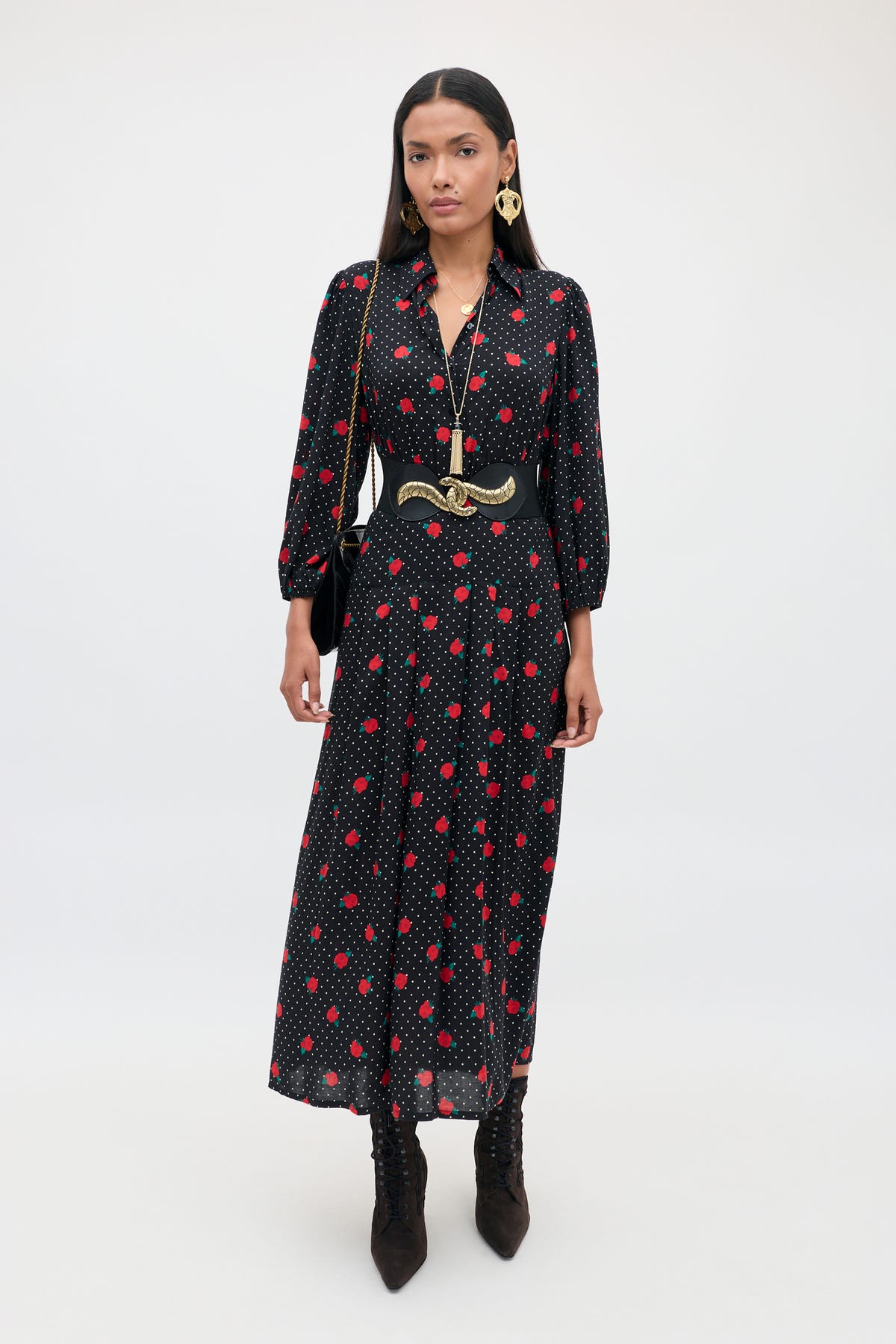 A woman stands against a plain background, wearing the Izzy - Rose Spot Black long dress with red polka dots, styled with black boots, a wide belt, gold jewelry, and carrying a black shoulder bag.