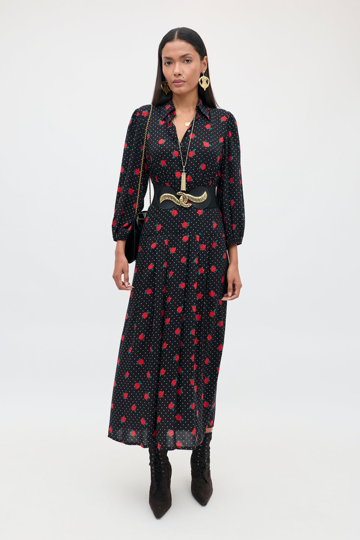 A woman stands against a plain background, wearing the Izzy - Rose Spot Black long dress with red polka dots, styled with black boots, a wide belt, gold jewelry, and carrying a black shoulder bag.