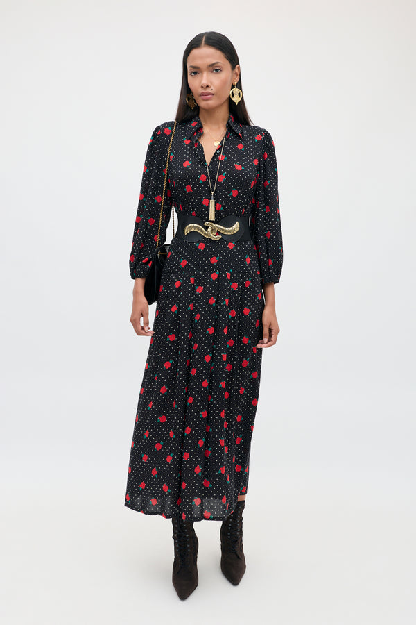 A woman stands against a plain background, wearing the Izzy - Rose Spot Black long dress with red polka dots, styled with black boots, a wide belt, gold jewelry, and carrying a black shoulder bag.