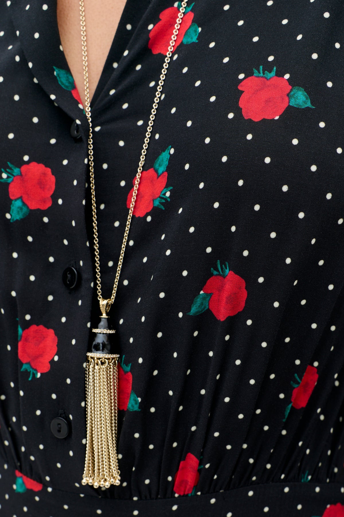 A close-up of the Izzy - Rose Spot Black dress, featuring a black fabric with white polka dots and red roses, styled with a gold tassel necklace adorned with a black accent.