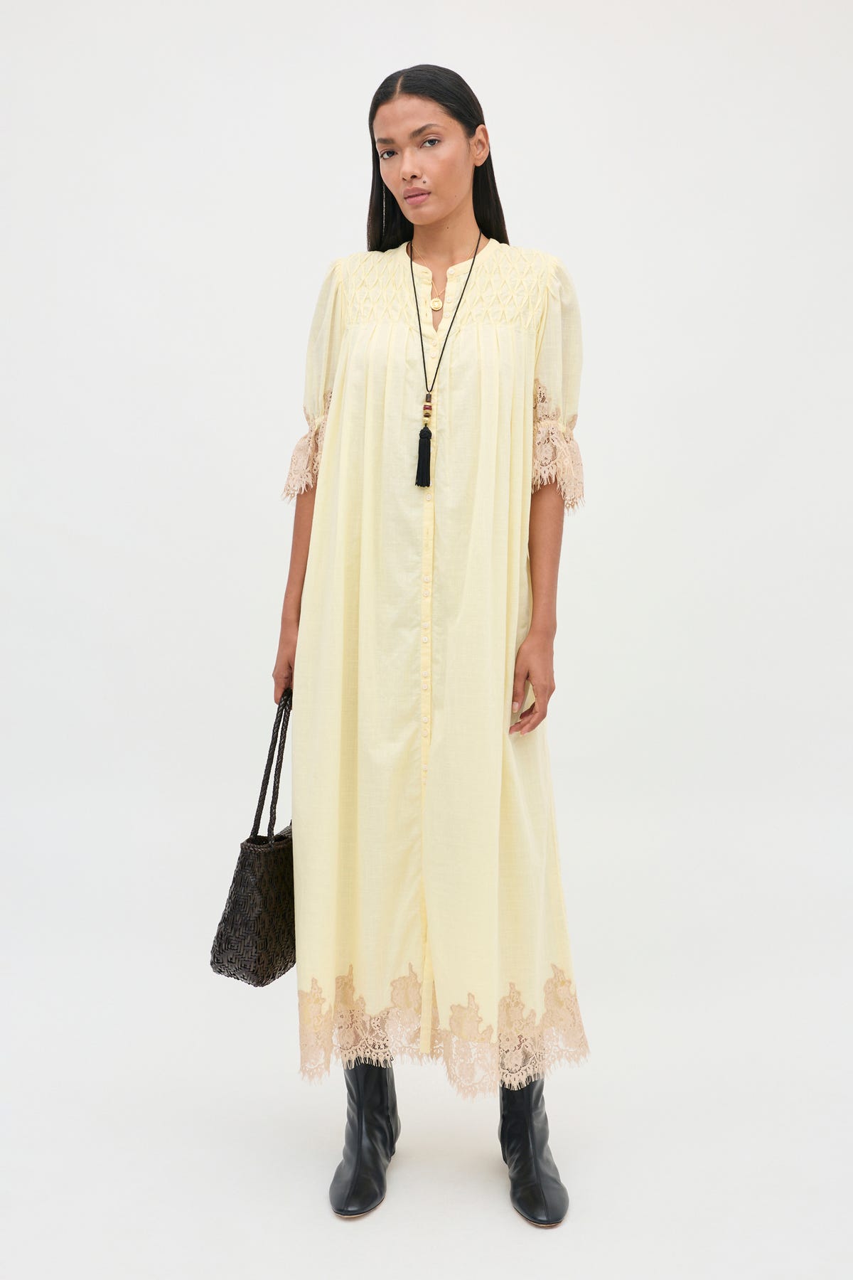 A woman wearing the Lorela - Lemon pale yellow, mid-calf length dress with lace trim on the sleeves and hem, paired with black boots and carrying a dark woven bag.