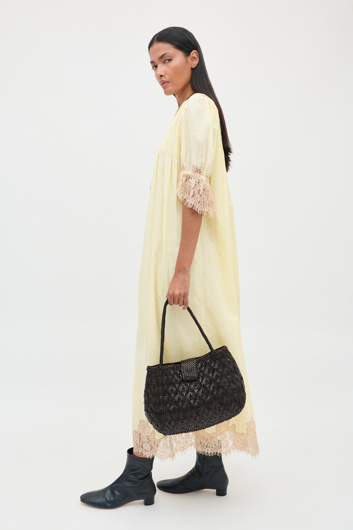 A person wears a Lorela - Lemon pale yellow midi dress with lace trim at the sleeves and hem, paired with black leather ankle boots and a dark brown woven handbag.