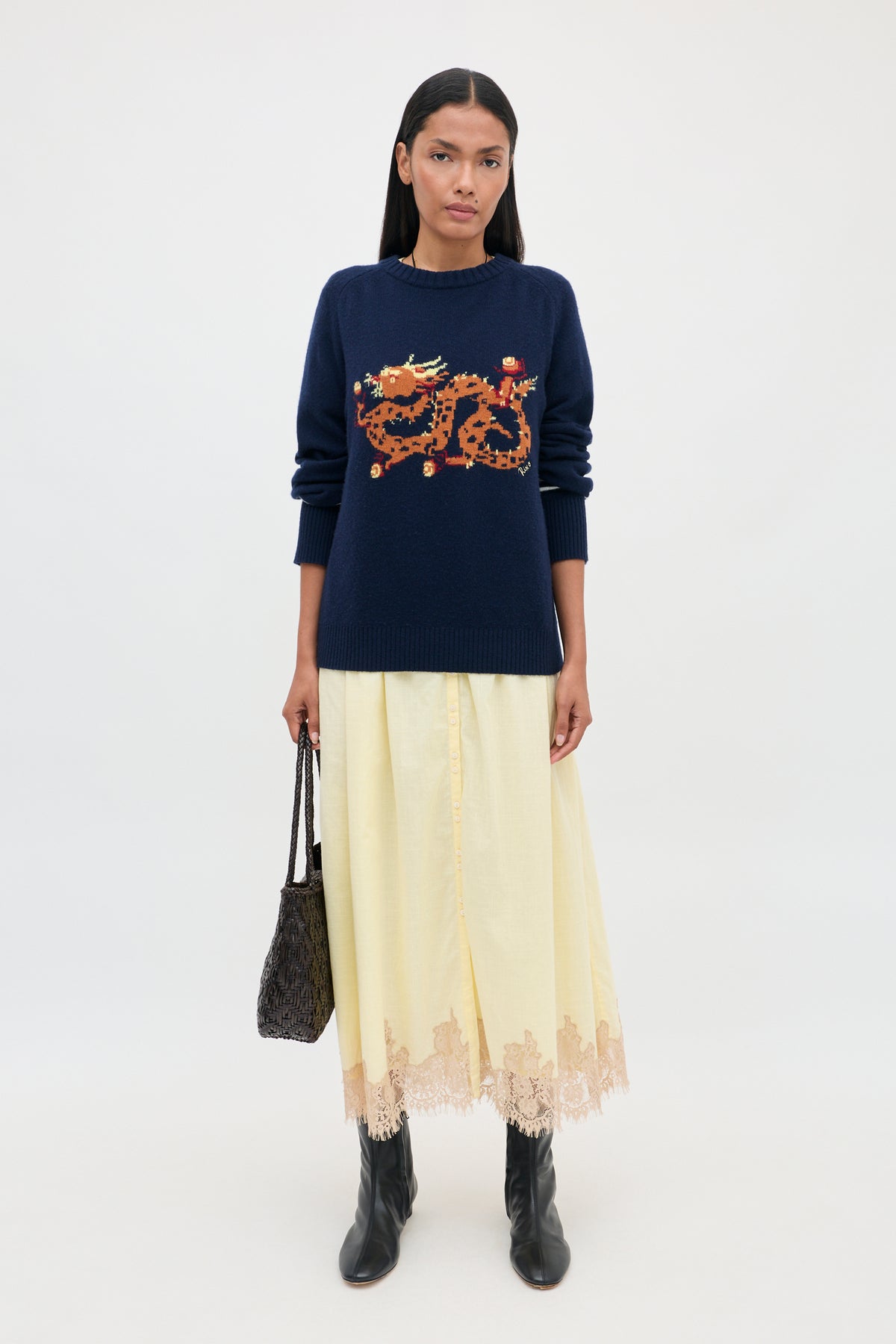 A person wears a navy blue crewneck sweater with a pixelated dragon graphic and the Lorela - Lemon skirt, which is long, pale yellow, and features lace trim, paired with black leather boots.