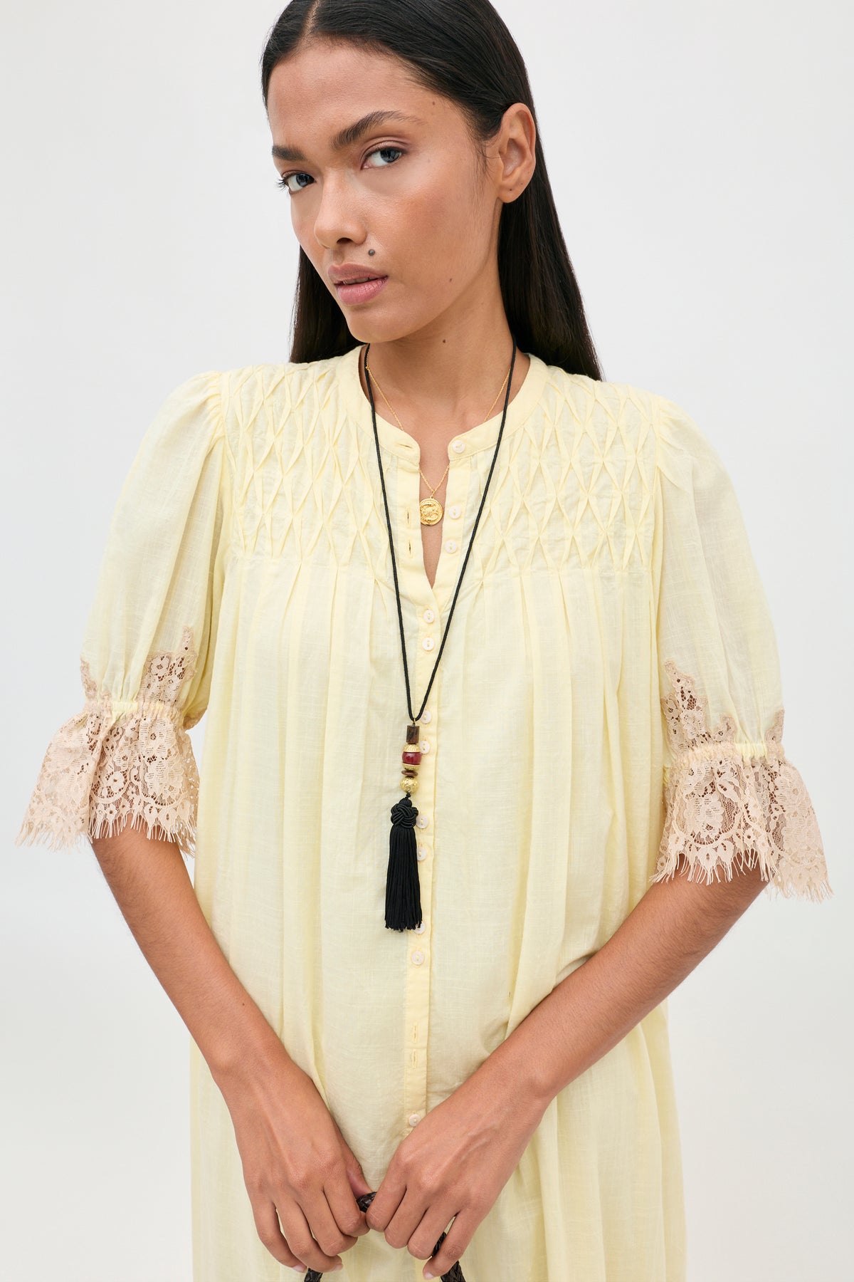 A person wearing the Lorela - Lemon pale yellow button-down blouse with gathered detailing and beige lace-trimmed sleeves, accessorized with two layered necklaces.