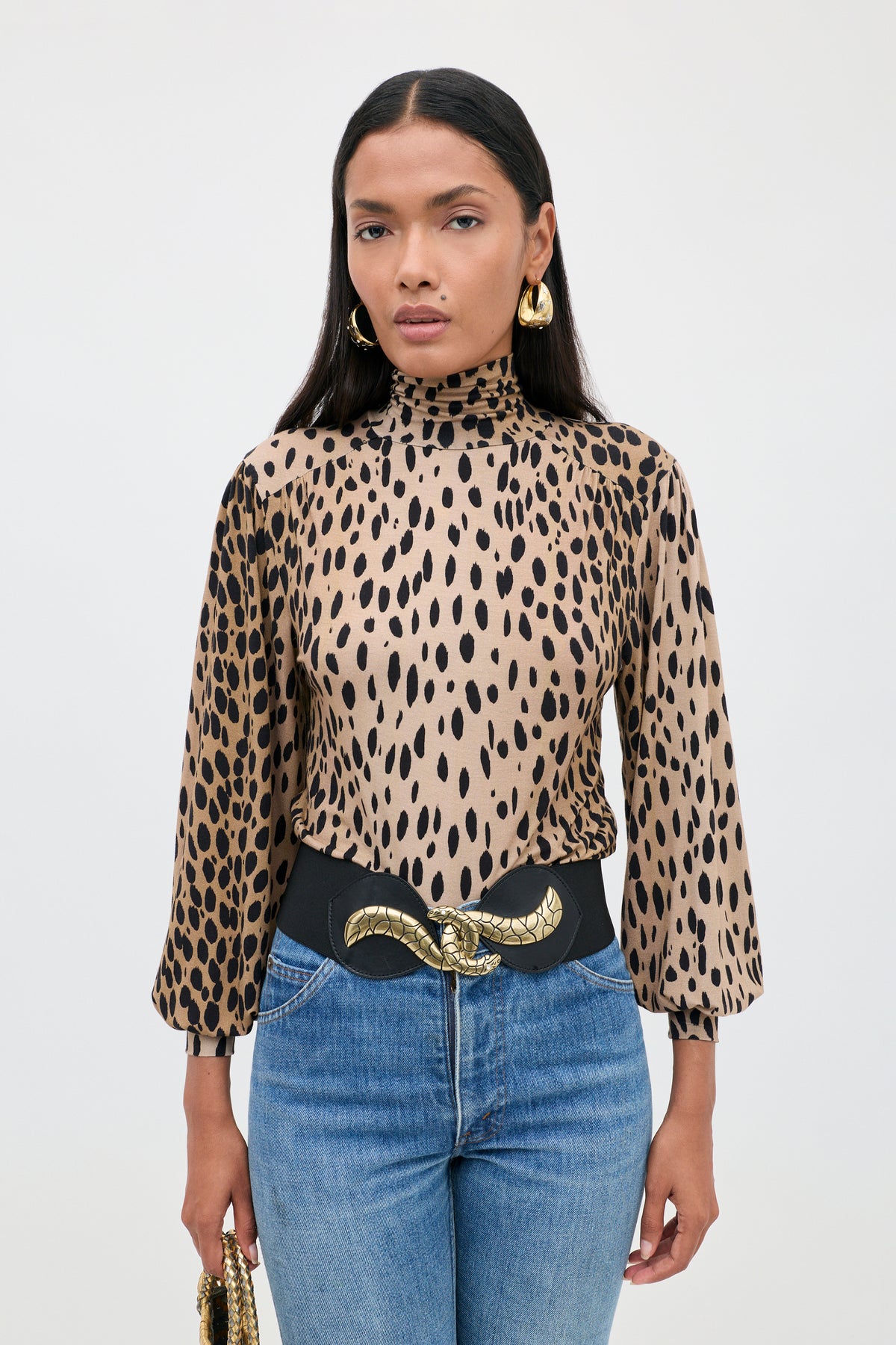 A woman wearing the Suki - Antelope Spot Black turtleneck blouse, gold hoop earrings, a black belt with a gold snake buckle, and blue jeans stands against a plain white background.