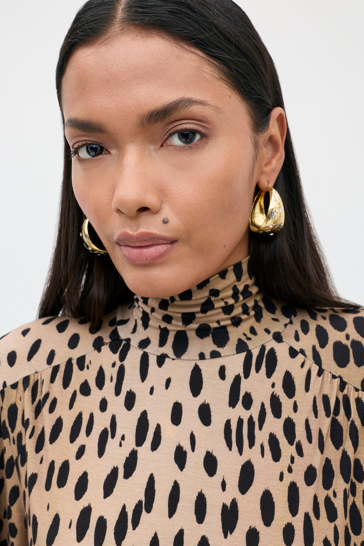A person with straight dark hair wears large gold hoop earrings and the Suki - Antelope Spot Black top, looking directly at the camera with a neutral expression.
